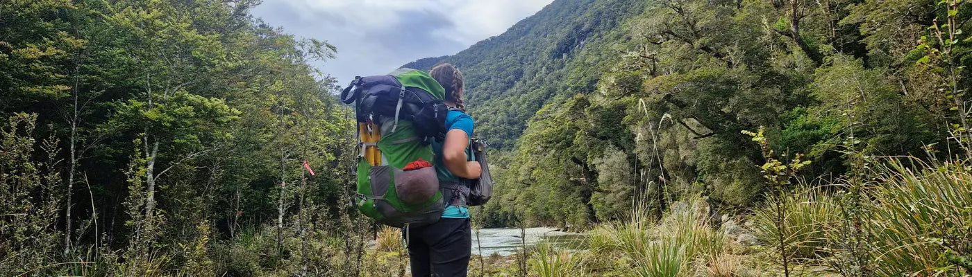 A woman is standing, looking at the view, next to a lake, in Fiordland National Park. She's hiking and packrafting, and wear an Aarn Peak aspiration. 