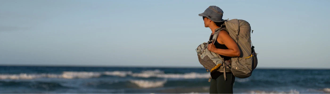 A woman is hiking on a beach, next to the ocean, with her Aarn hiking pack.
