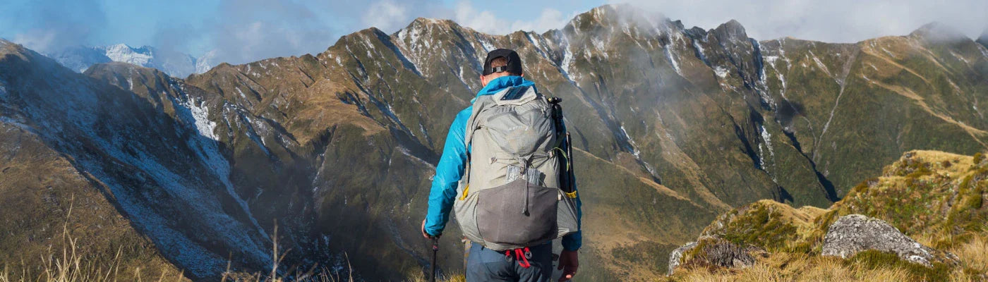 A man is standing on top of a mountain, looking at the view. He is carrying a grey lightweight tramping pack.